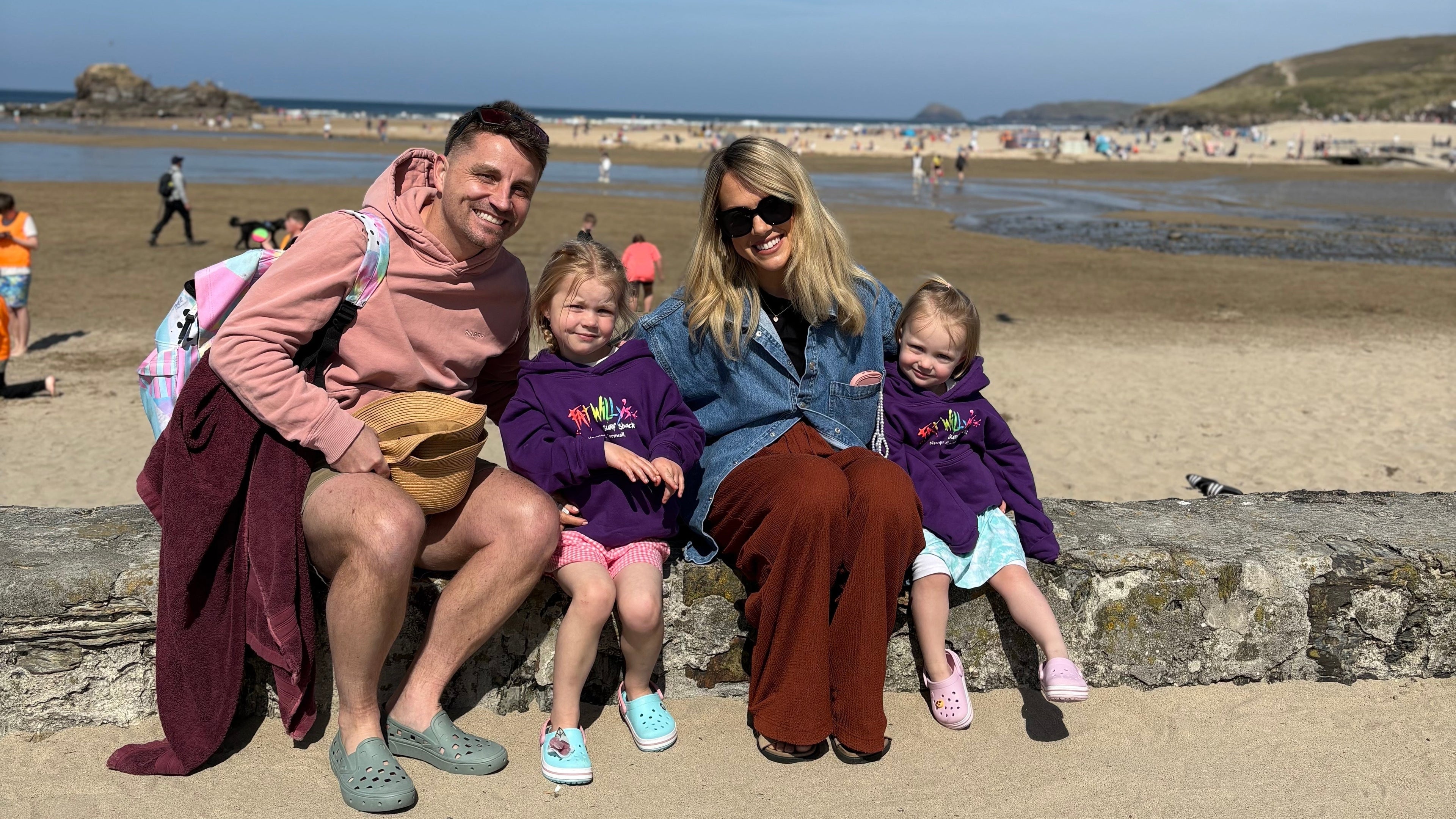 Family of four sitting on a stone ledge by a beach with a clear blue sky.