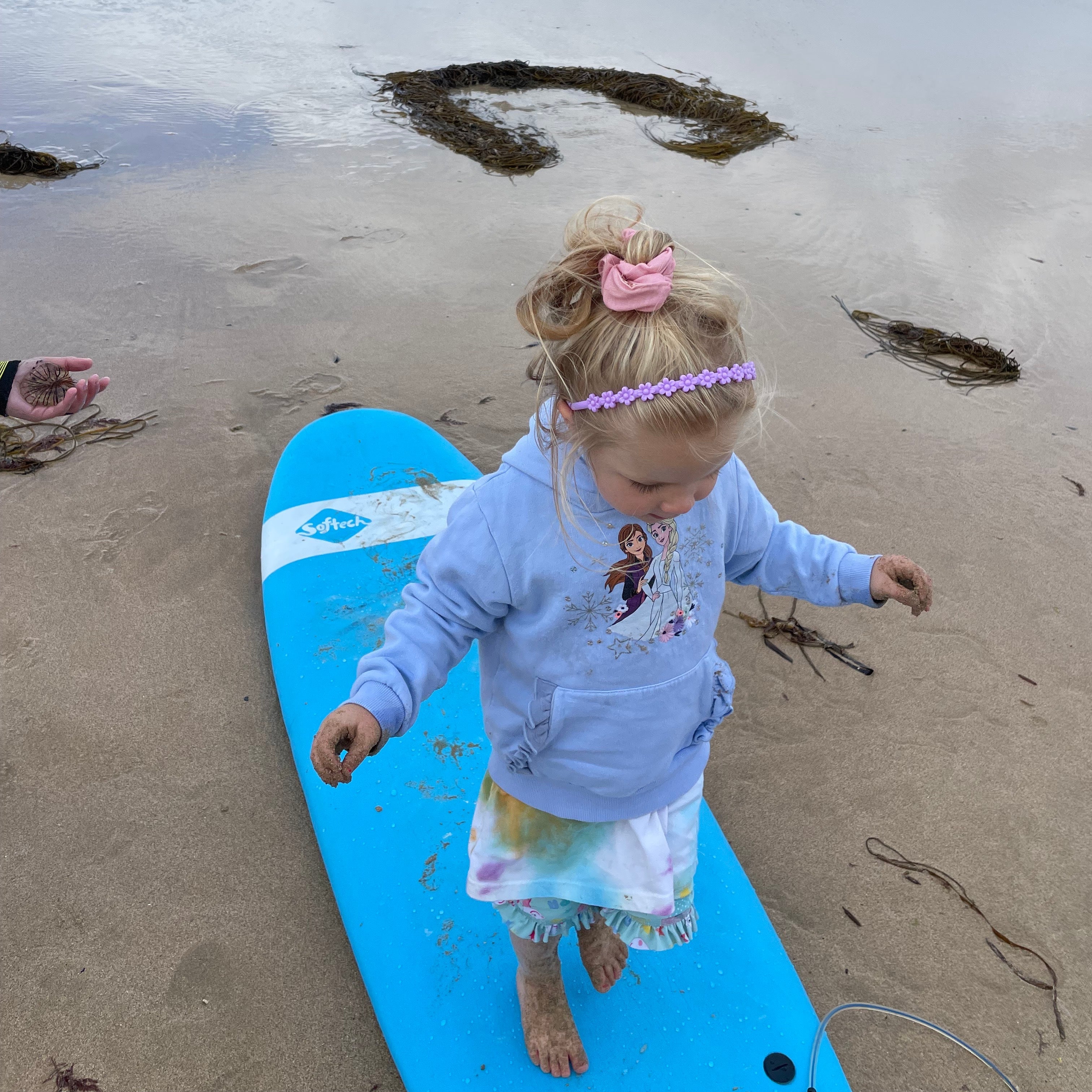Child standing on a blue surfboard at the beach with ocean and sky in the background